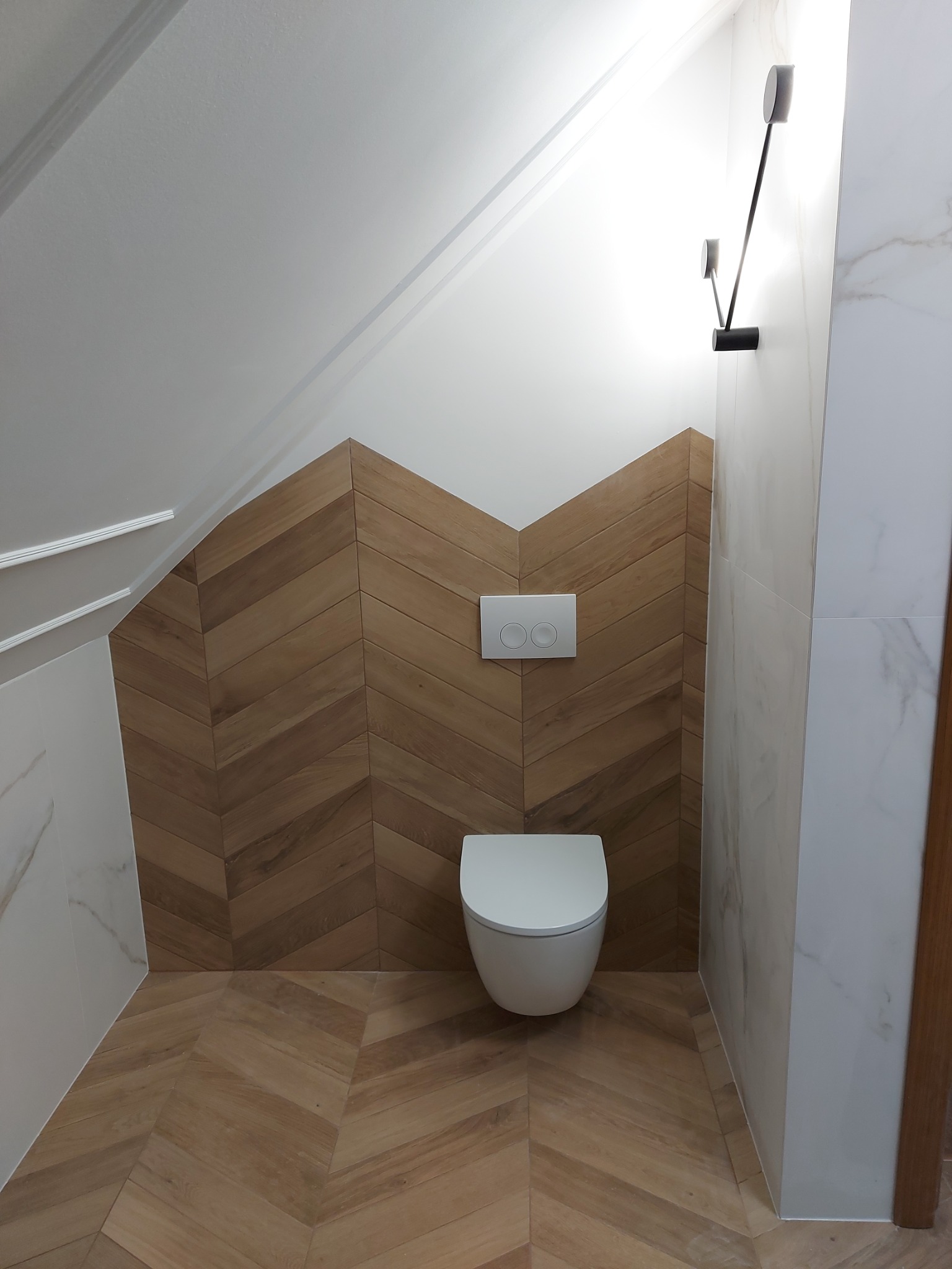 Nowoczesna toaleta w skosach: drewniana mozaika na ścianie i podłodze ułożona w jodełkę, biała ceramika sanitarna, minimalistyczna lampa ścienna.