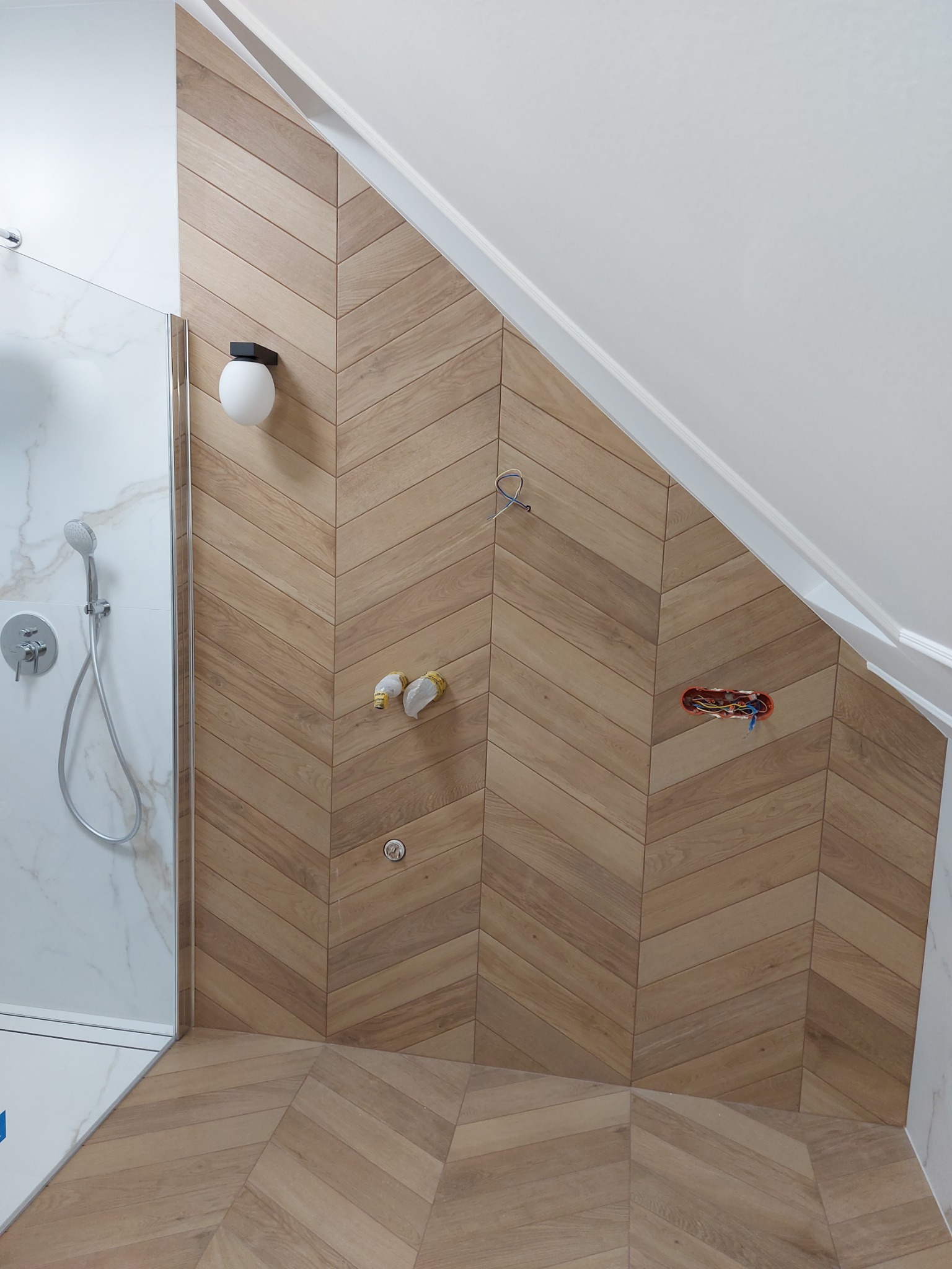 Nowoczesna łazienka na poddaszu z drewnopodobnymi płytkami ułożonymi w jodełkę na ścianie i podłodze. Widoczna kabina prysznicowa i przygotowane punkty instalacyjne.