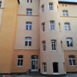 Odnowiona fasada budynku z charakterystyczną okrągłą wieżą, pomalowana na odcienie beżu i pomarańczy, widoczne okna z białymi ramami i fragment placu z materiałami budowlanymi.