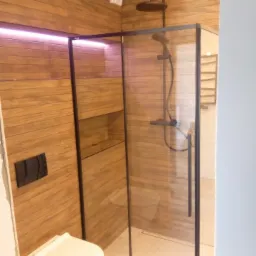 Nowoczesna łazienka z kabiną prysznicową typu walk-in z czarnymi profilami, drewnopodobnymi płytkami na ścianach, podświetleniem LED i wiszącą miską WC.