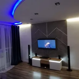 Nowoczesny salon z podświetlanym sufitem LED w kolorze niebieskim, dekoracyjną ścianą z motywem chevron, telewizorem i szafką RTV z oświetleniem LED.