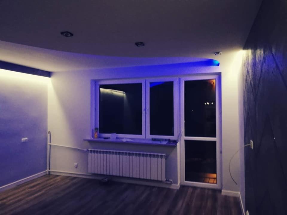 Pokój po renowacji z podwieszanym sufitem z oświetleniem LED w kolorze niebieskim, drewniana podłoga i ściana z dekoracyjną tapetą w odcieniach szarości.