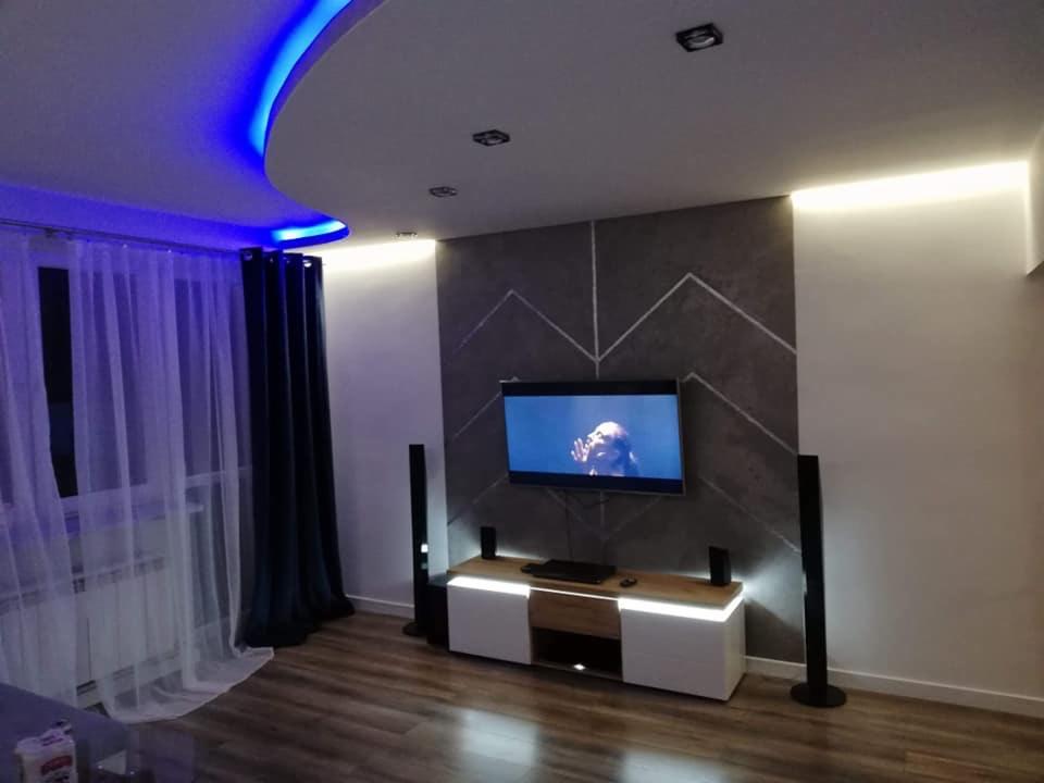 Nowoczesny salon z podświetlanym sufitem LED w kolorze niebieskim, dekoracyjną ścianą z motywem chevron, telewizorem i szafką RTV z oświetleniem LED.