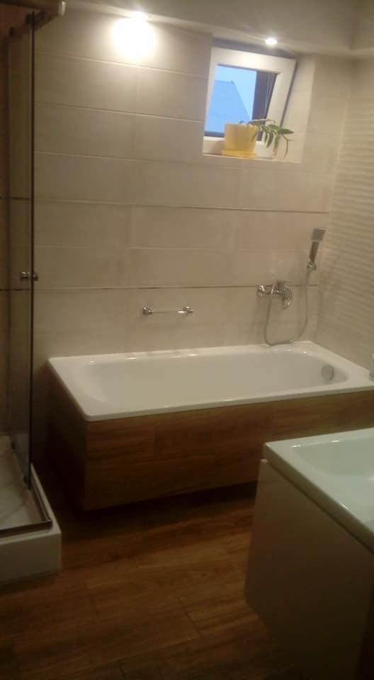 Nowoczesna łazienka z wanną obudowaną drewnem, kabiną prysznicową ze szklanymi drzwiami i drewnianą podłogą; na ścianie jasne płytki i okno z rośliną w żółtej doniczce.