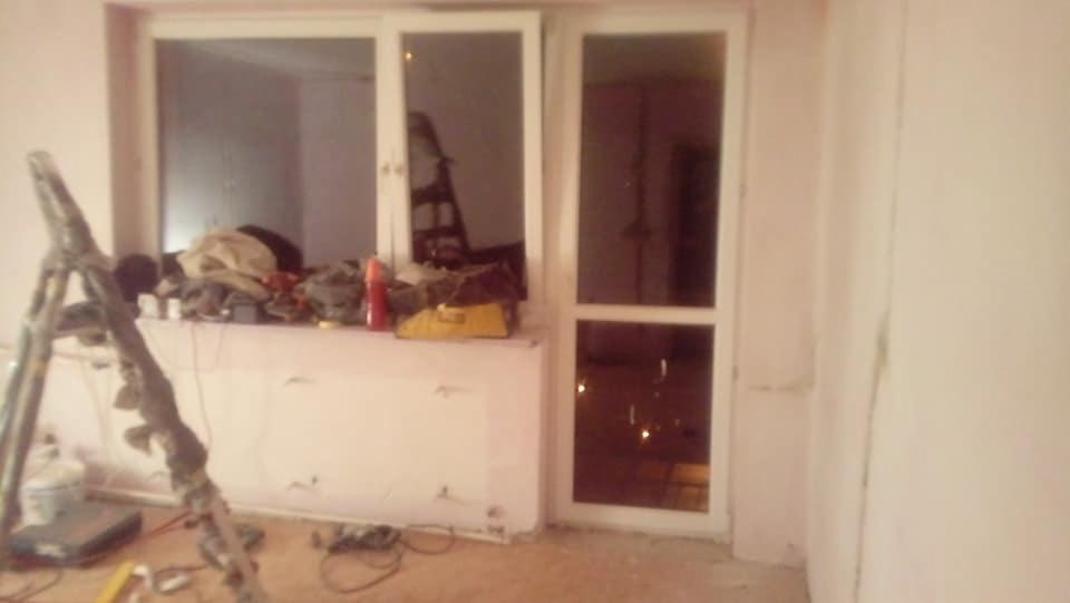 Wnętrze w trakcie prac wykończeniowych: białe okno z drzwiami balkonowymi, drabina malarska oparta o ścianę, narzędzia i materiały budowlane na parapecie, ściany w jasnym odcieniu różu.