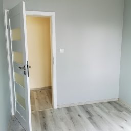 Mar-Bud - Świeżo pomalowane szare ściany w pokoju z otwartymi białymi drzwiami z przeszkleniami i panelami podłogowymi w odcieniach szarości, widoczny przełącznik światła na ścianie.