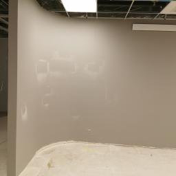 Mar-Bud - Ściana w trakcie przygotowania do malowania, widoczne ślady po szpachlowaniu i gruntowaniu, surowy sufit z instalacjami, zaokrąglony narożnik ściany.