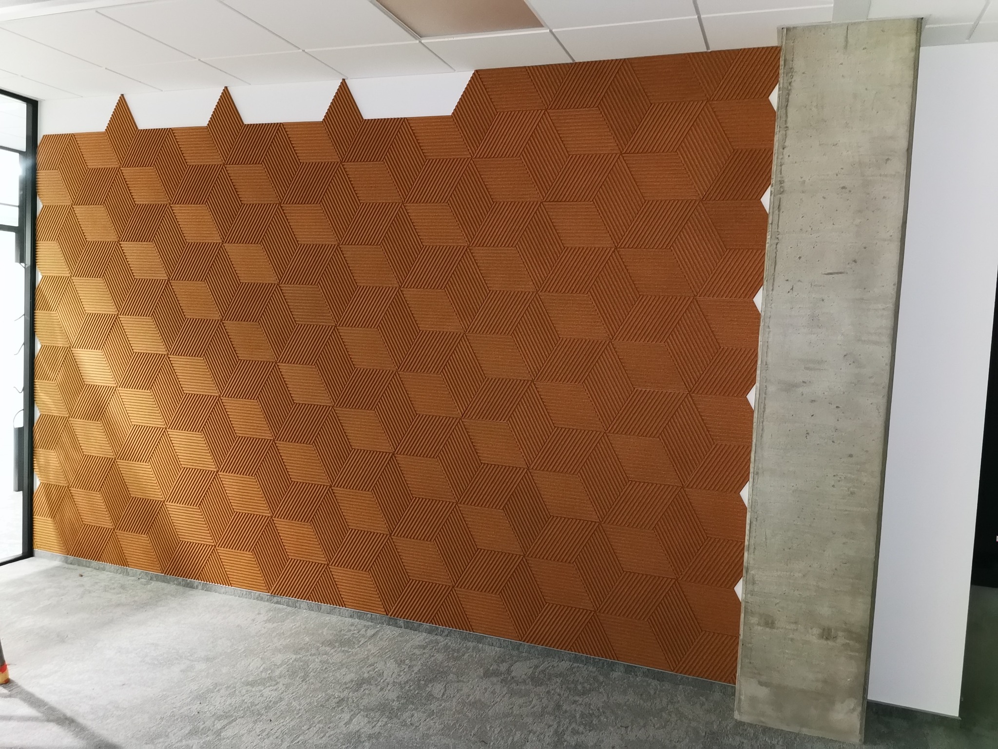 Wykończona ściana z dekoracyjnymi panelami 3D w odcieniach brązu, tworzącymi geometryczny wzór kostek, widoczny betonowy filar i fragment sufitu z niedokończonymi fragmentami.