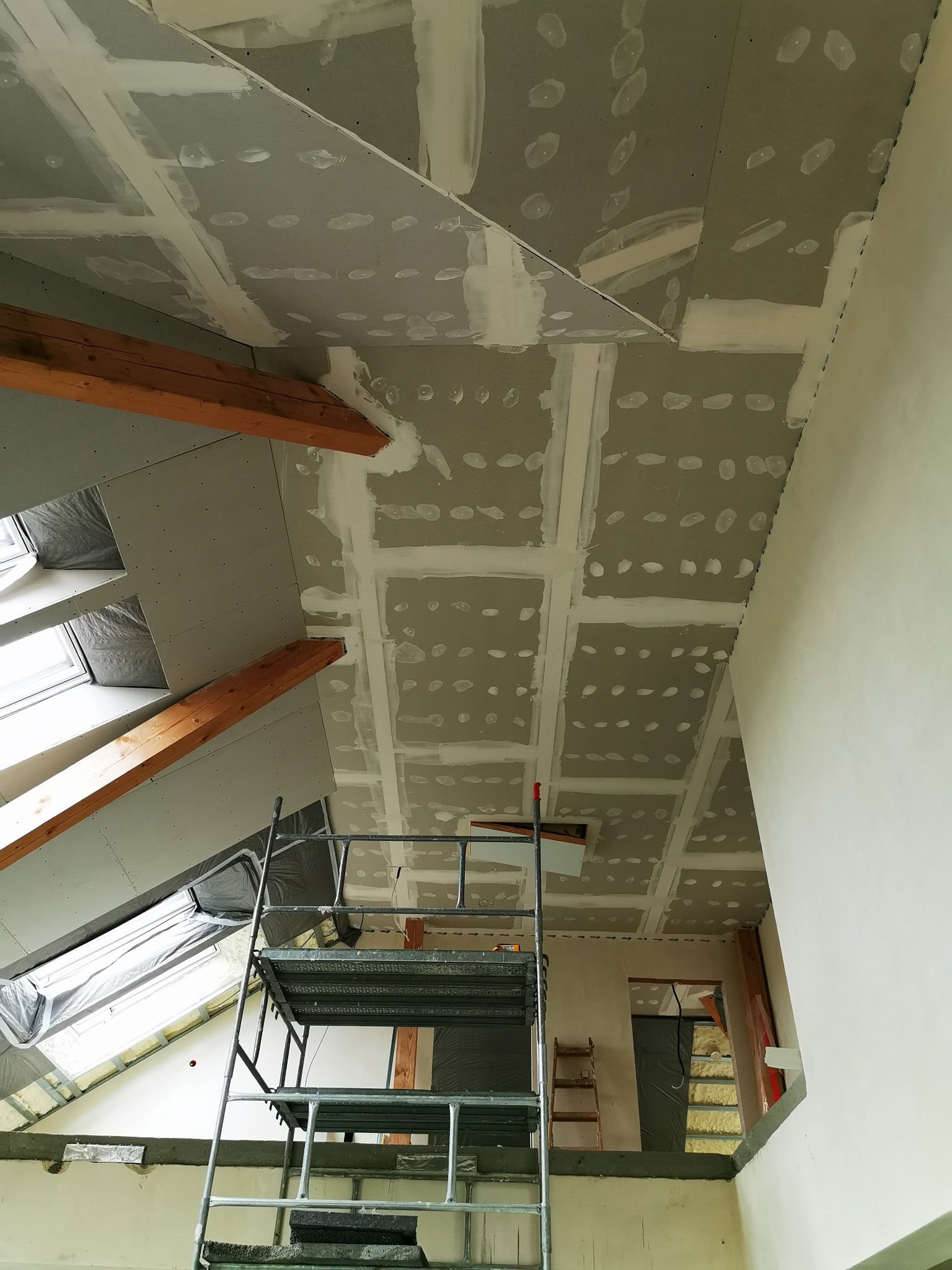 Skośny sufit poddasza z zamontowanymi płytami gipsowo-kartonowymi, widoczne drewniane belki konstrukcyjne i częściowo zaszpachlowane łączenia, z boku okno dachowe, na pierwszym planie metalowy ruszt.