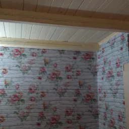 Ściana wykończona tapetą imitującą białą cegłę z motywem róż, widoczny fragment drewnianego sufitu i narożnik szafy.