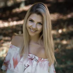 Portret uśmiechniętej, młodej kobiety z blond włosami w bluzce z motywem magnolii na tle rozmytej zieleni i brązu.