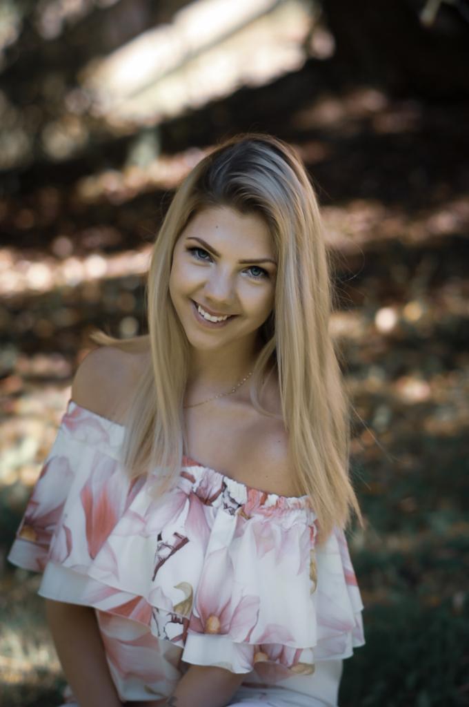 Portret uśmiechniętej, młodej kobiety z blond włosami w bluzce z motywem magnolii na tle rozmytej zieleni i brązu.