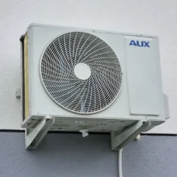 Zewnętrzna jednostka klimatyzacji AUX zamontowana na ścianie budynku z elewacją w dwóch odcieniach szarości i bieli.