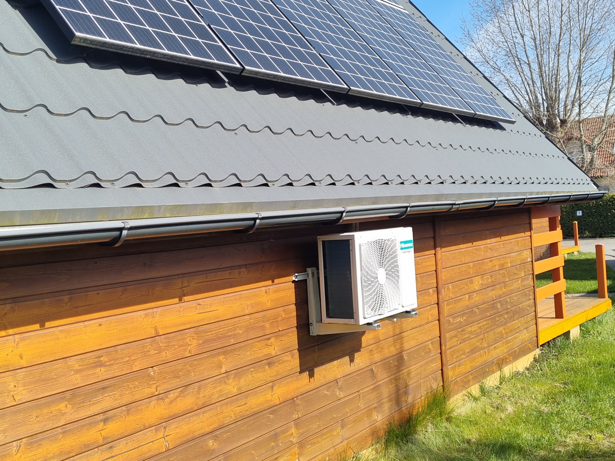 Boczny widok klimatyzatora Hisense zamontowanego na drewnianej ścianie domku z panelami słonecznymi na dachu.