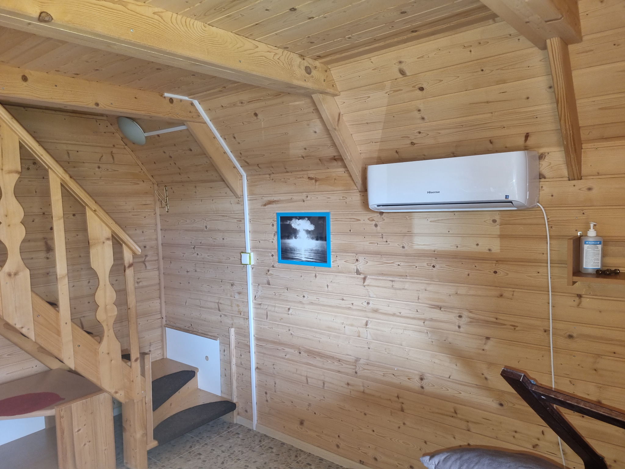 Wnętrze drewnianego domu z zamontowanym klimatyzatorem Hisense na ścianie, widoczne schody i obrazek w niebieskiej ramce.