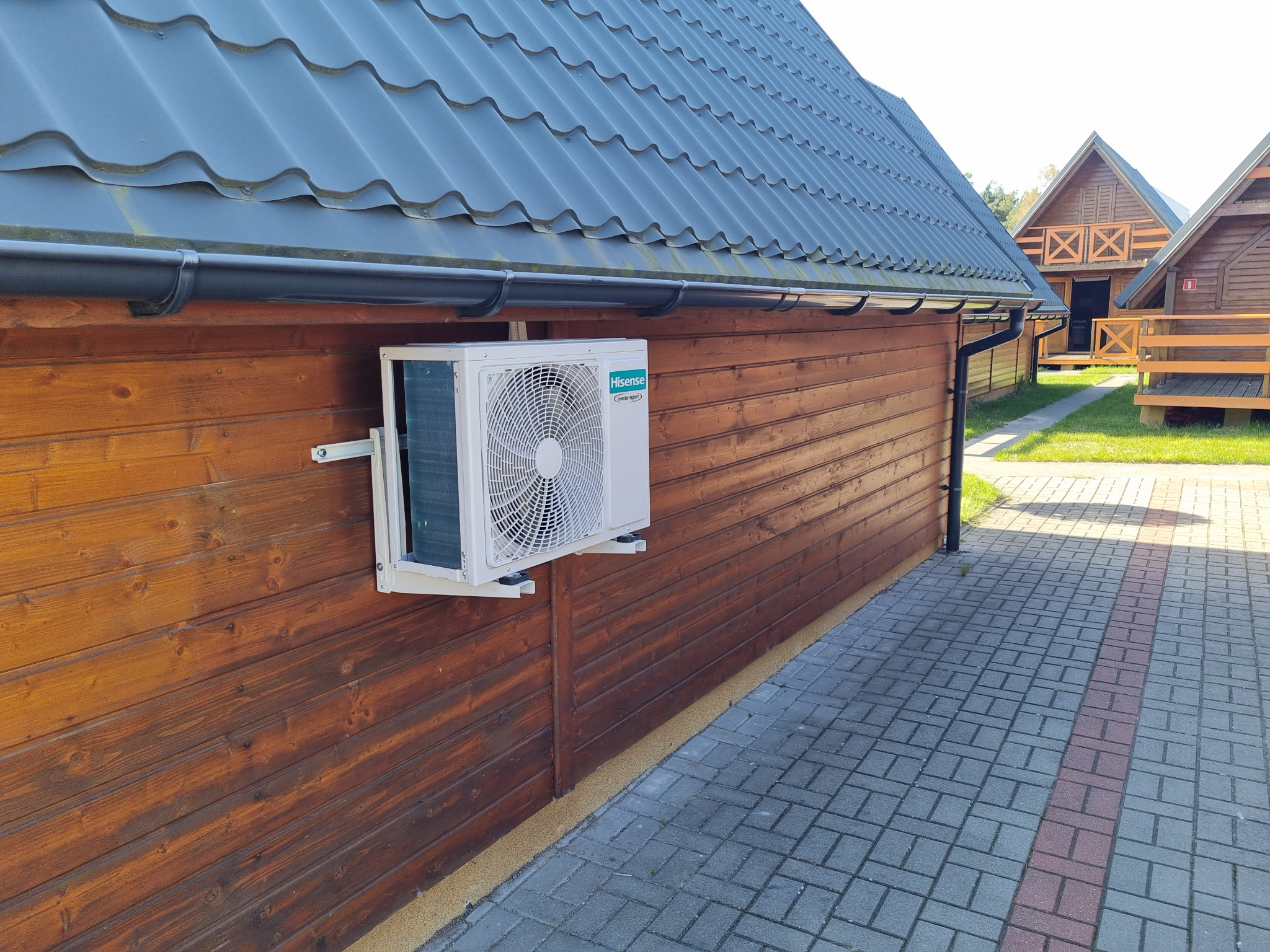 Zewnętrzna jednostka klimatyzacji Hisense zamontowana na drewnianej ścianie domku letniskowego z szarym dachem, widok z poziomu chodnika z kostki brukowej.