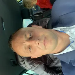 Mężczyzna w samochodzie ubrany w marynarkę, z dzieckiem widocznym w tle na tylnym siedzeniu.