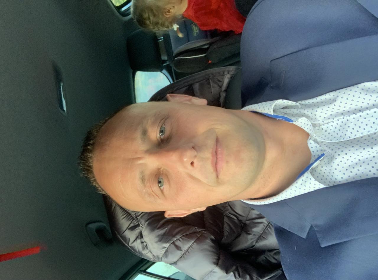 Mężczyzna w samochodzie ubrany w marynarkę, z dzieckiem widocznym w tle na tylnym siedzeniu.