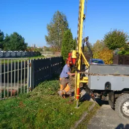 Pracownik w pomarańczowych spodniach obsługuje żółtą wiertnicę zamontowaną na samochodzie ciężarowym, wykonując odwiert w gruncie przy metalowym ogrodzeniu na tle błękitnego nieba i drzew.