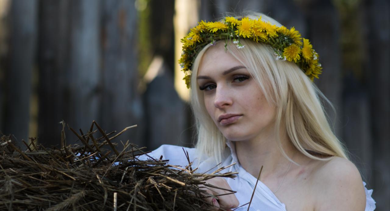 Portret młodej kobiety z blond włosami i wiankiem z żółtych dmuchawców na głowie, trzymającej w rękach wiązkę suchych gałęzi na tle drewnianego ogrodzenia.