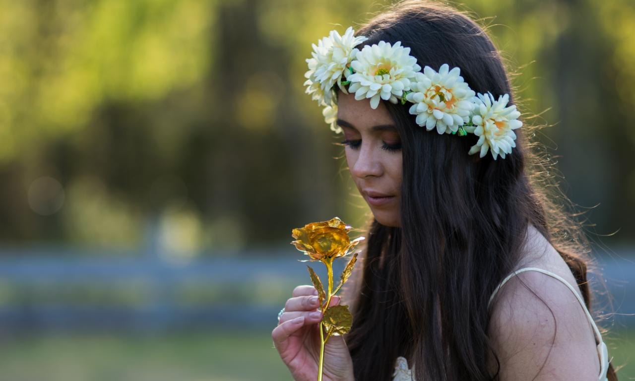 Kobieta z wiankiem na głowie, trzymająca złotą różę w dłoni, spogląda w dół, z zamazanym zielonym tłem.