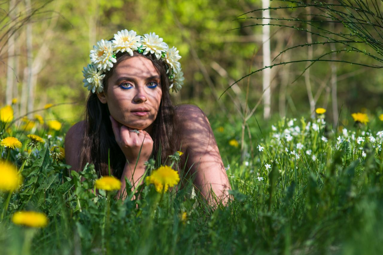 Kobieta z ciemnymi włosami i kwiatowym wiankiem na głowie, leżąca na łące pełnej żółtych i białych kwiatów, z cieniem padającym na twarz.