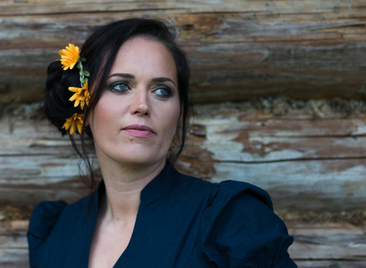 Portret kobiety z ciemnymi włosami upiętymi z żółtymi kwiatami, na tle drewnianej ściany.