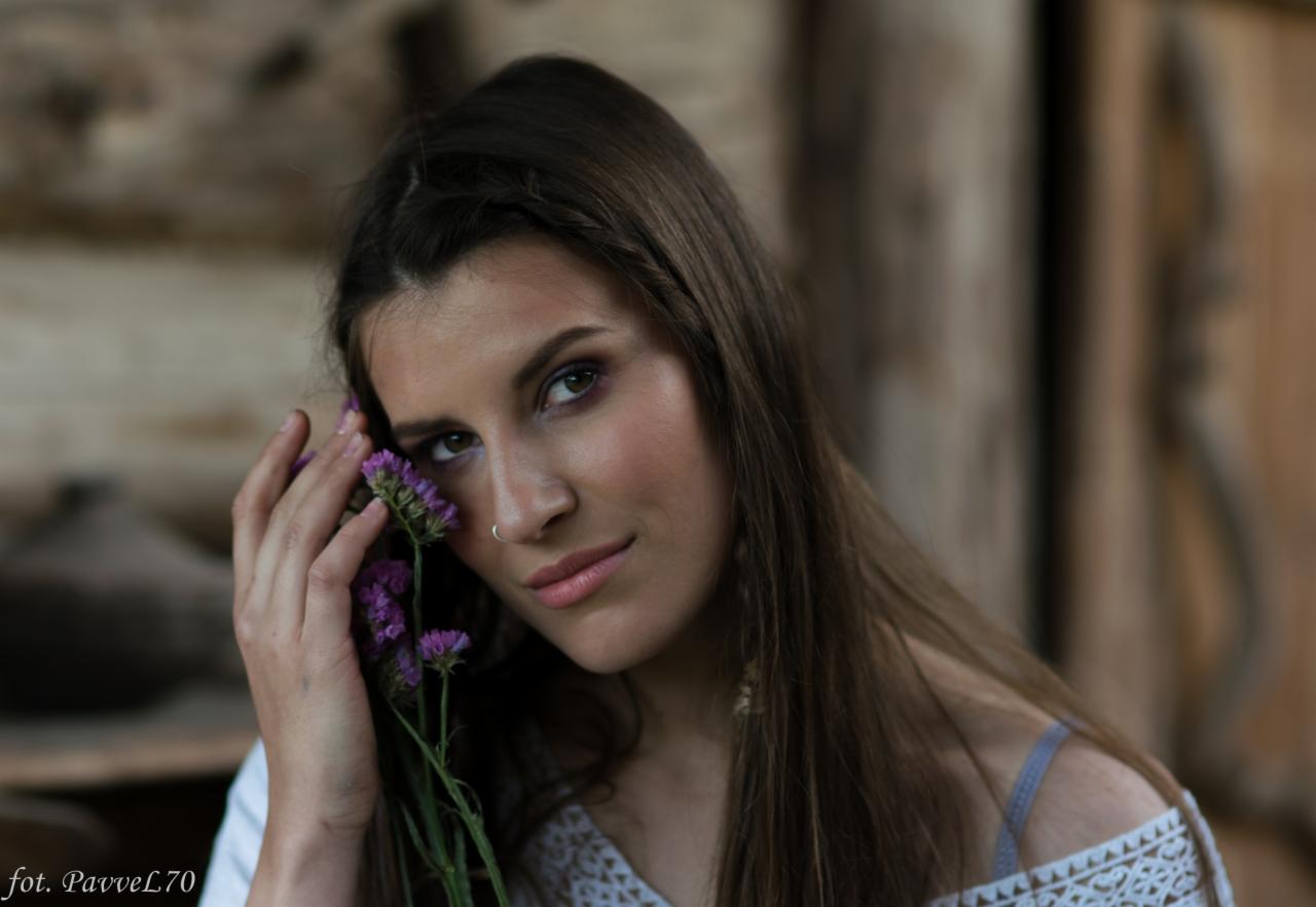 Portret młodej kobiety z kolczykiem w nosie, trzymającej gałązkę fioletowych kwiatów przy twarzy, na tle drewnianej ściany.