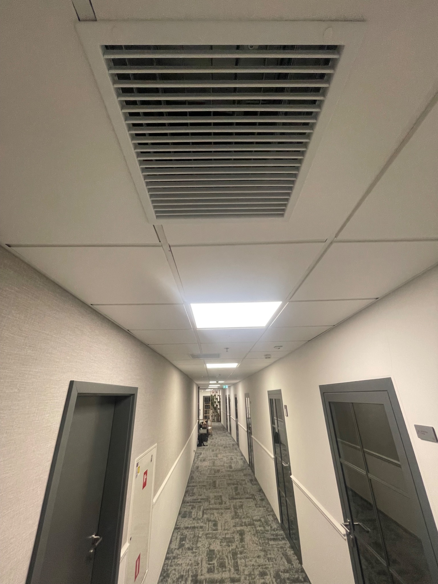 Sufitowy system wentylacyjny z kratką w korytarzu budynku komercyjnego, widok z dołu na długi korytarz z drzwiami po obu stronach.