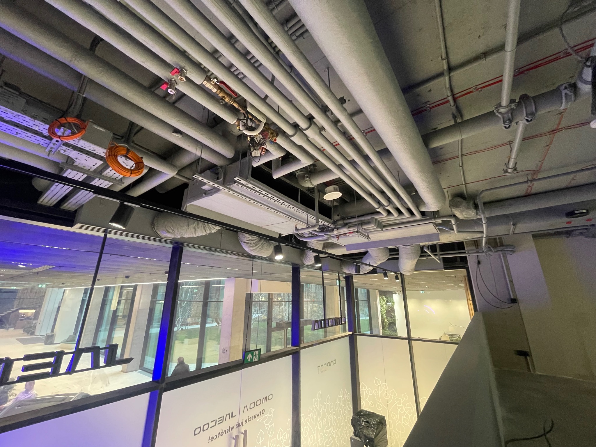 Szereg rur wentylacyjnych pod sufitem w biurowcu w Warszawie, widoczne kable i elementy mocujące, z zewnątrz widok na budynek przez szklaną ścianę.