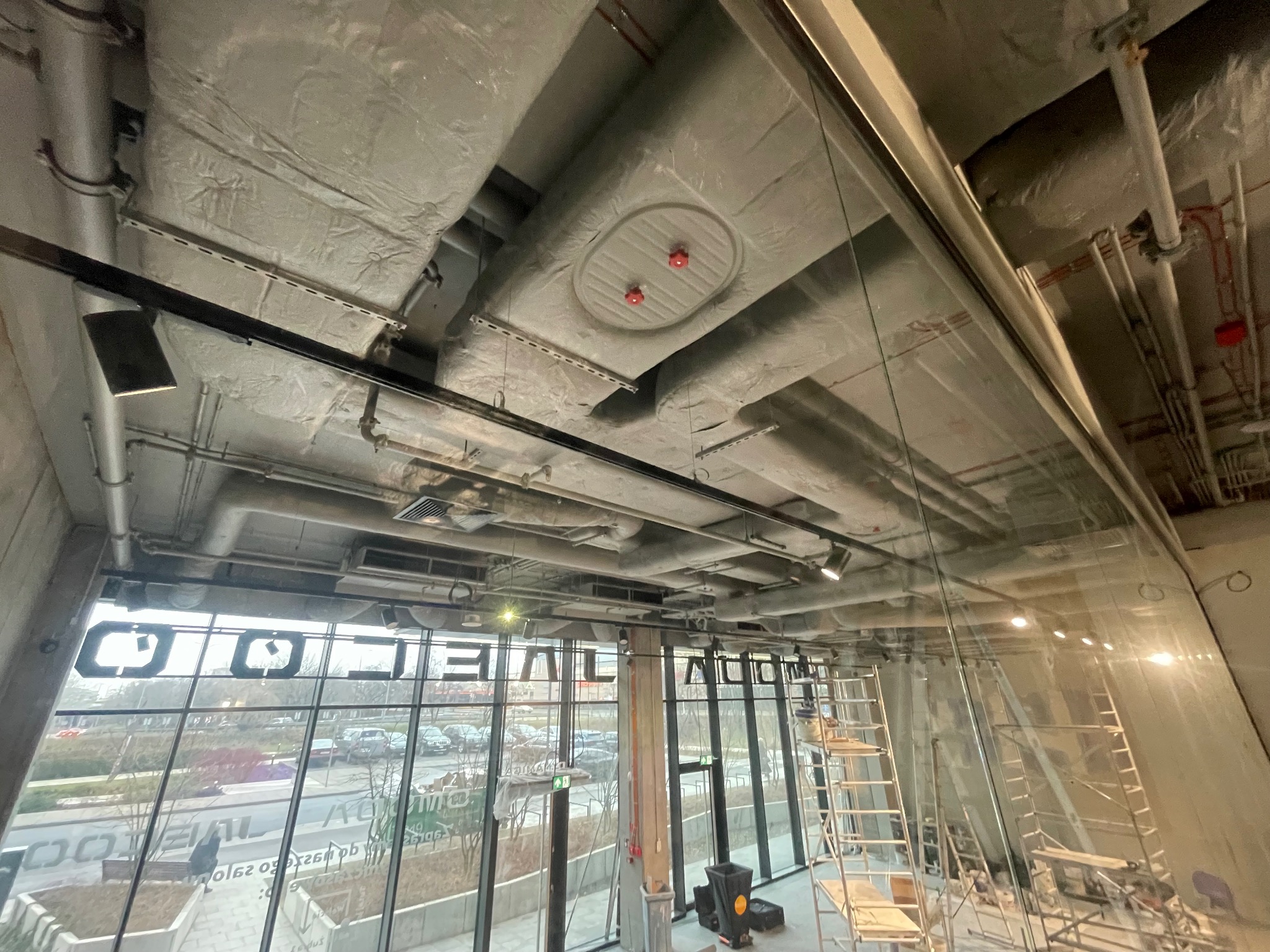 Widok na sufit budynku komercyjnego z widocznymi przewodami wentylacyjnymi w izolacji, elementami instalacji przeciwpożarowej oraz widokiem na zewnątrz przez duże okna.