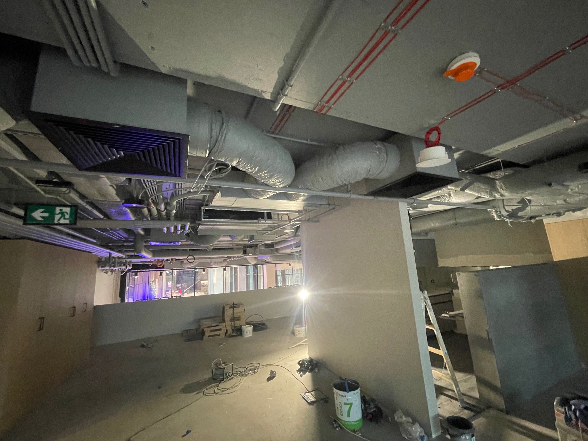 Wnętrze budynku komercyjnego w trakcie prac wykończeniowych; widoczne rury wentylacyjne pod sufitem, instalacje elektryczne, drabina oparta o filar i puszki z farbą na podłodze