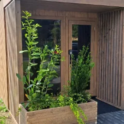 Nowoczesna, drewniana konstrukcja z przeszklonymi drzwiami, otoczona roślinami w drewnianej skrzyni i wyłożona kostką brukową.