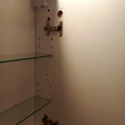 Wnętrze białej szafki z zamontowanymi zawiasami i półkami szklanymi, widoczne ślady po wierceniu otworów.