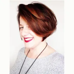 Uśmiechnięta kobieta z krótkimi, brązowymi włosami z rudymi refleksami, czerwonymi ustami i czarnym naszyjnikiem na tle białej ściany.