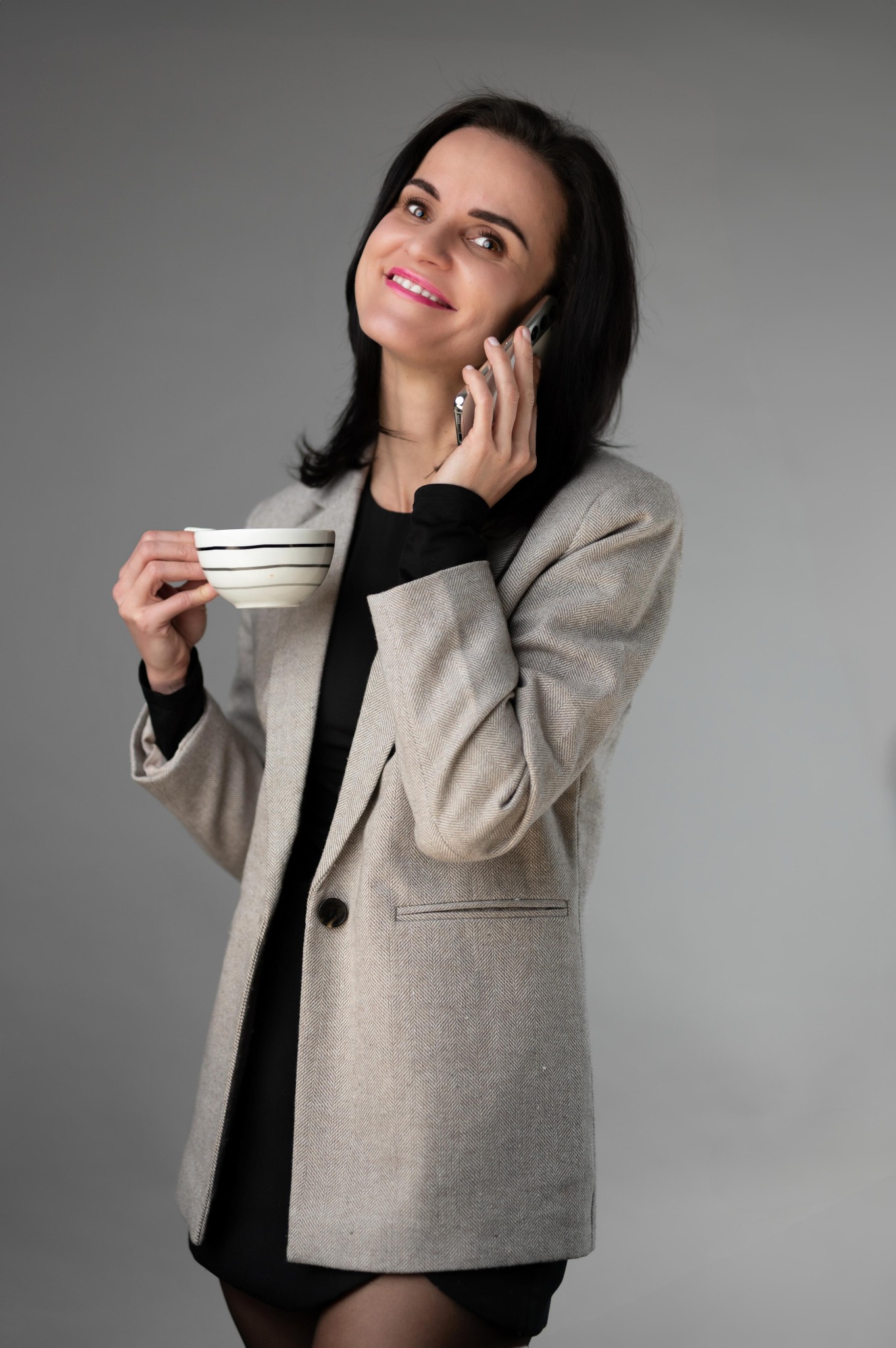 Uśmiechnięta brunetka w tweedowej marynarce rozmawia przez telefon, trzymając filiżankę w paski. Stonowane tło, profesjonalny wizerunek.