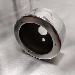 Polerowany, cylindryczny element metalowy z otworami na śruby, leżący na metalowej powierzchni z widocznymi śladami obróbki.