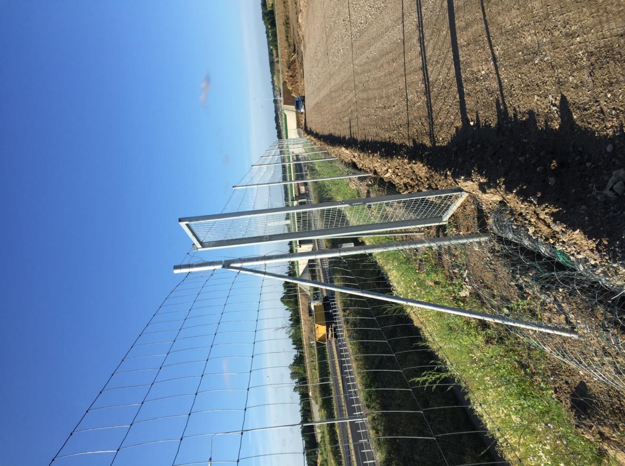 Montaż siatki ogrodzeniowej wzdłuż drogi, widoczne metalowe słupki i częściowo rozciągnięta siatka, świeżo usypana ziemia po wykonanych pracach.