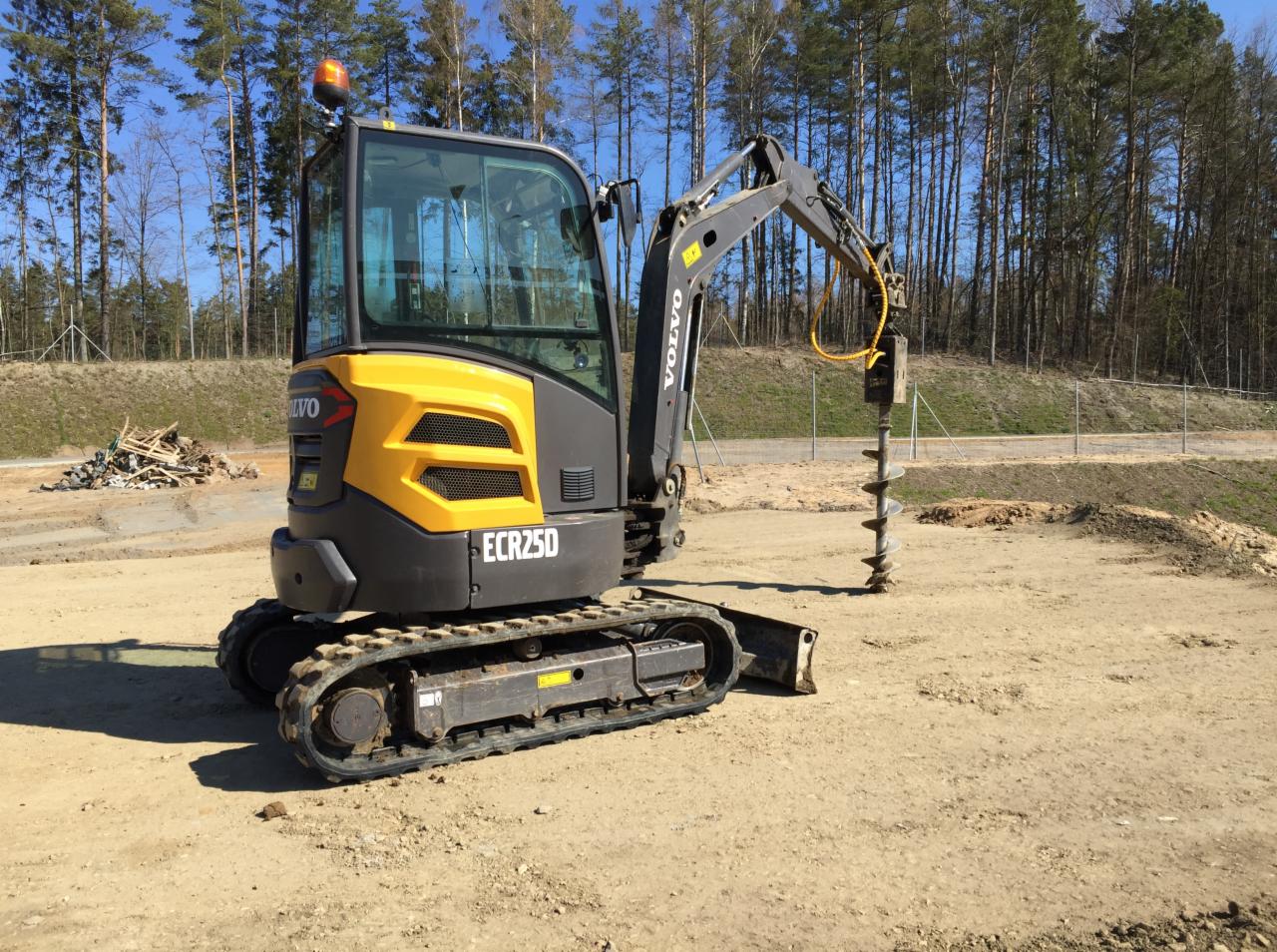 Żółto-szara minikoparka Volvo ECR25D z wiertnicą na placu budowy, w tle las i stos gruzu. Słoneczny dzień, błękitne niebo.