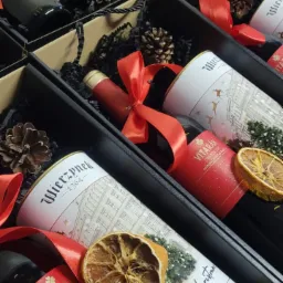 Świąteczne kosze prezentowe z winem i piernikami Wierzynek, zapakowane w czarne pudełka z dekoracjami w Bielsko-Białej. Czerwone wstążki i suszone pomarańcze.