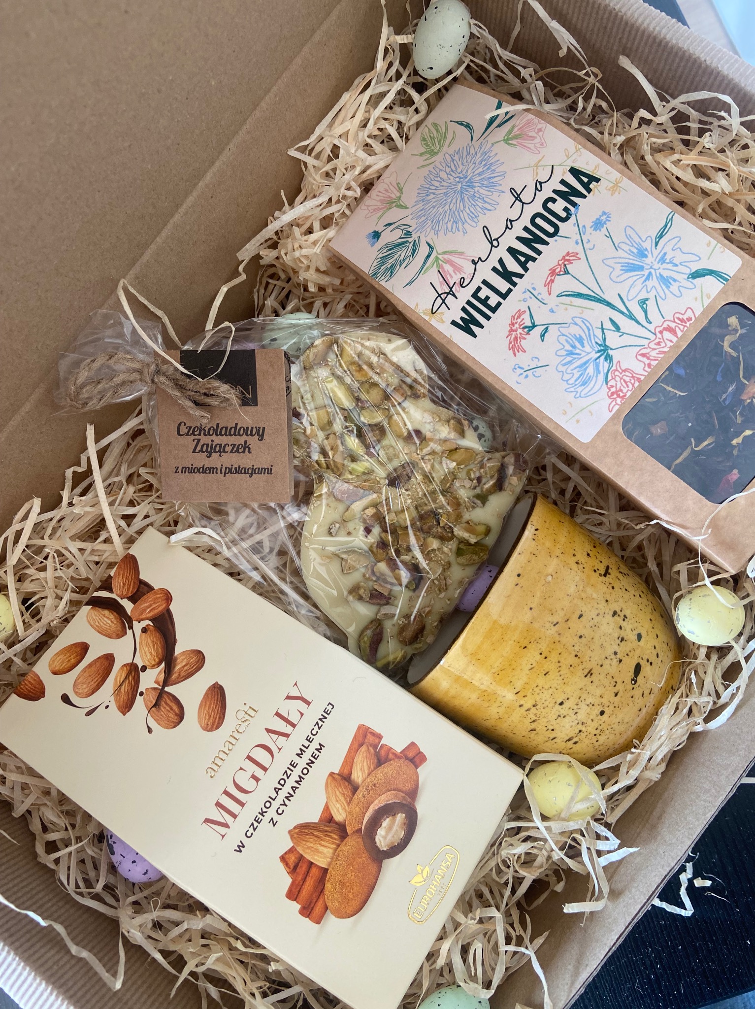 Otwarty kartonowy box wypełniony siankiem, zawierający zestaw prezentowy: czekoladowy zajączek z miodem i pistacjami w folii, pudełko herbaty wielkanocnej z kwiatowym motywem, tabliczka czekolady...