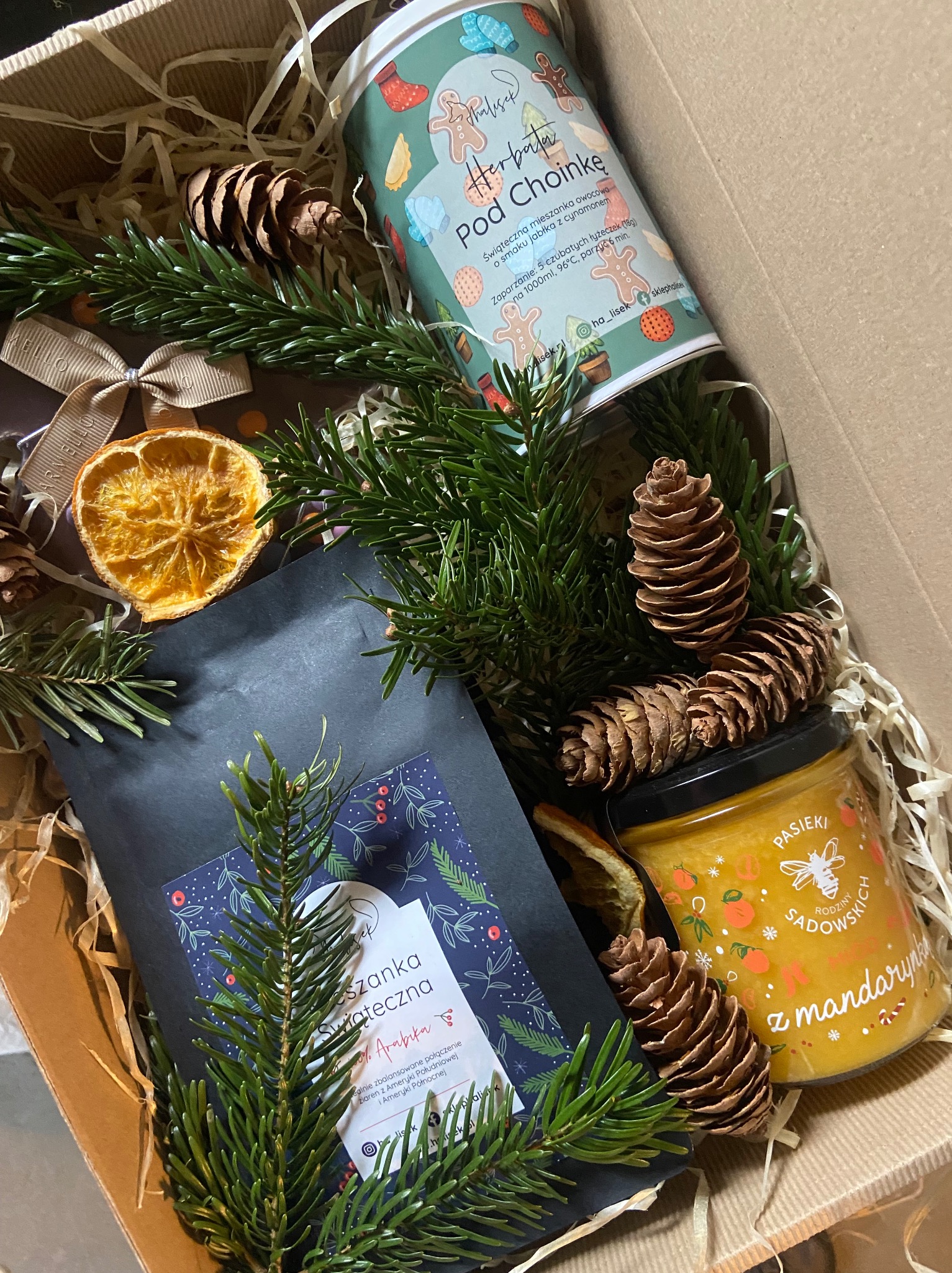 Świąteczny zestaw upominkowy z lokalnymi produktami od firmy z Bielska-Białej, zawierający herbatę w puszce, kawę w czarnym opakowaniu i słoik miodu z mandarynkami, udekorowany suszoną pomarańczą...