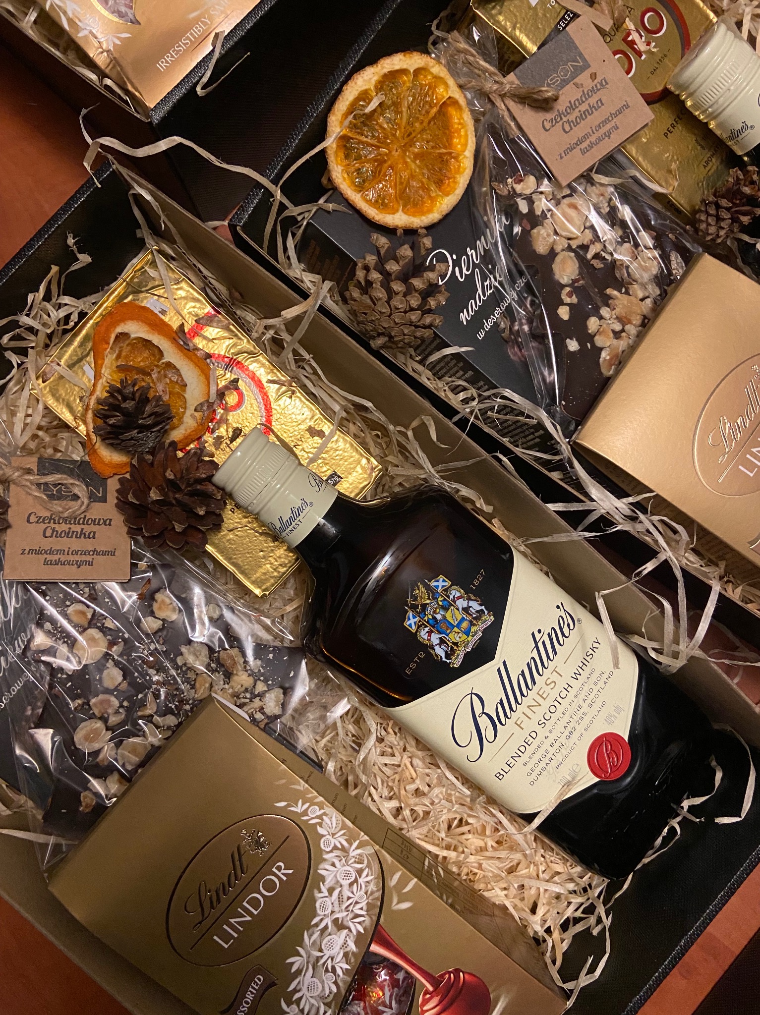 Elegancki czarny kartonowy box wypełniony whisky Ballantine's, złotymi pralinami Lindt, czekoladą z orzechami laskowymi, suszonymi pomarańczami i szyszkami na dekoracyjnym sianku.