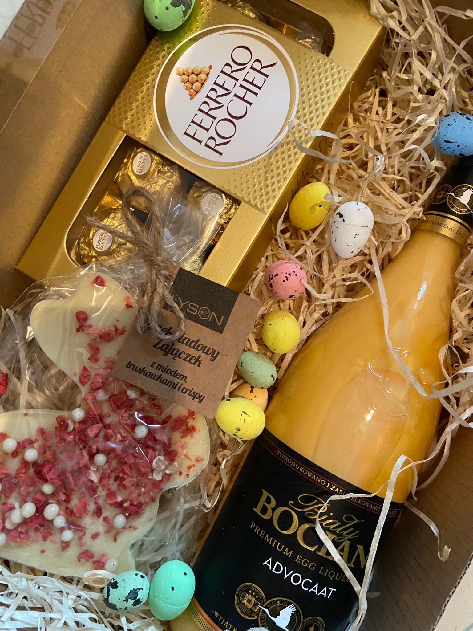 Świąteczny kosz prezentowy z czekoladkami Ferrero Rocher, likierem Advocat, czekoladowym zajączkiem i dekoracyjnymi jajkami w brązowym pudełku wypełnionym siankiem.