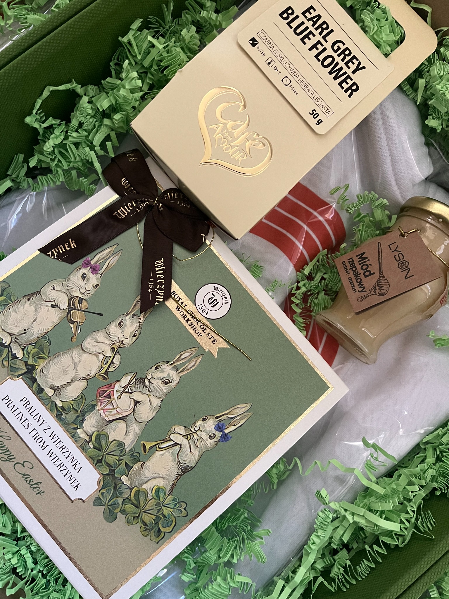 Elegancki kosz prezentowy z pralinami Wierzynek, herbatą Earl Grey Blue Flower i miodem rzepakowym Łysoń, wypełniony zielonym siankiem dekoracyjnym.