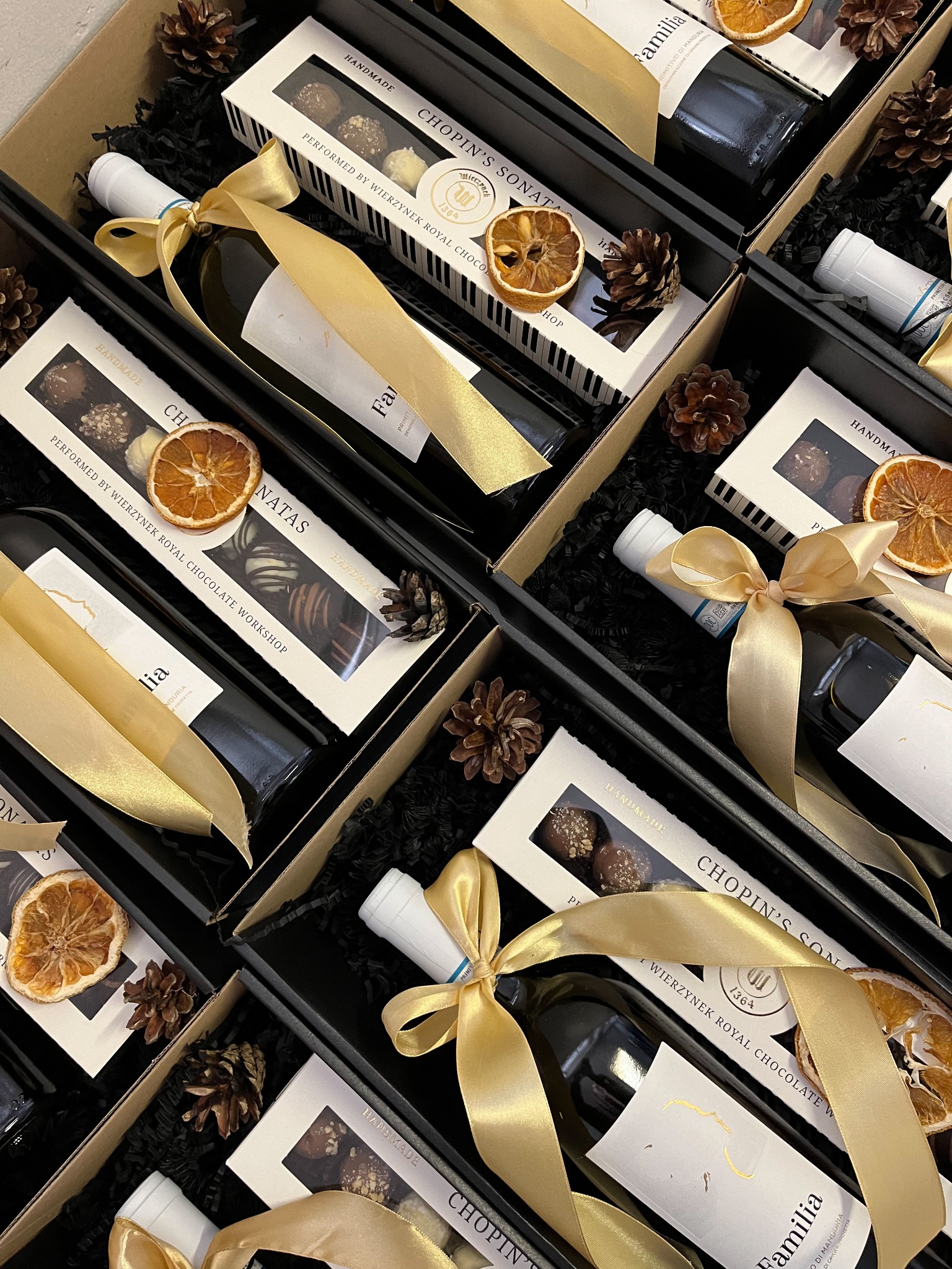 Eleganckie kosze prezentowe z winem, pralinkami i dekoracjami, zapakowane w czarne pudełka, przewiązane złotymi wstążkami. Widoczne szyszki i suszone pomarańcze.