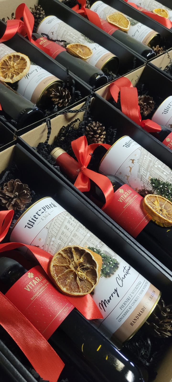 Świąteczne kosze prezentowe z winem i piernikami Wierzynek, zapakowane w czarne pudełka z dekoracjami w Bielsko-Białej. Czerwone wstążki i suszone pomarańcze.