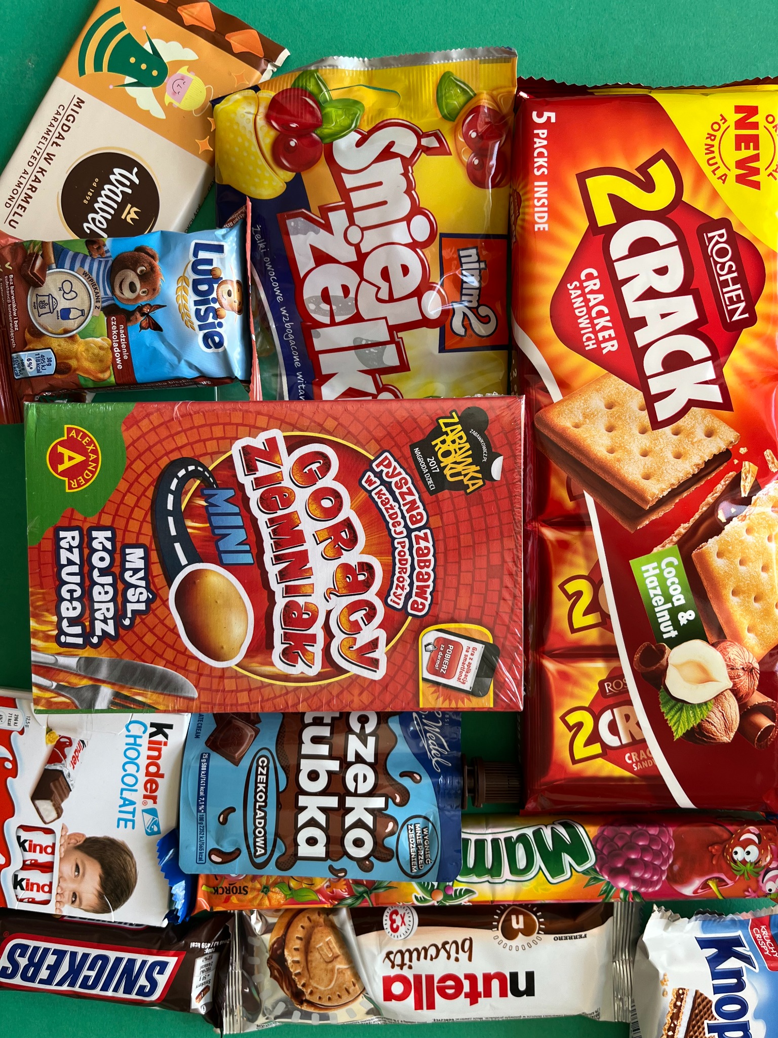 Słodki asortyment: czekolady Kinder, Snickers, Lubisie, żelki Śmiejżelki, wafle Knoppers, ciastka Nutella Biscuits i gra Gorący Ziemniak Mini na zielonym tle.