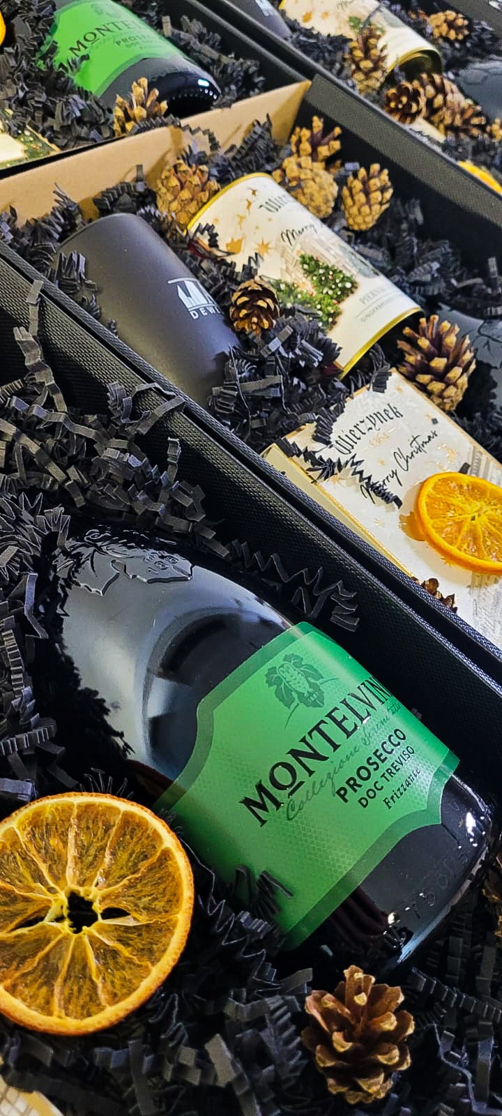 Elegancki kosz prezentowy z butelką prosecco Montelvin, czarną wyściółką, suszonymi pomarańczami i szyszkami, idealny na świąteczny upominek firmowy.