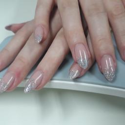 Studio Kosmetyczne La Mariposa - Dłonie kobiety z przedłużonymi, owalnymi paznokciami w kolorze nude z srebrnym brokatem na końcach, ułożone na szarym podłożu.